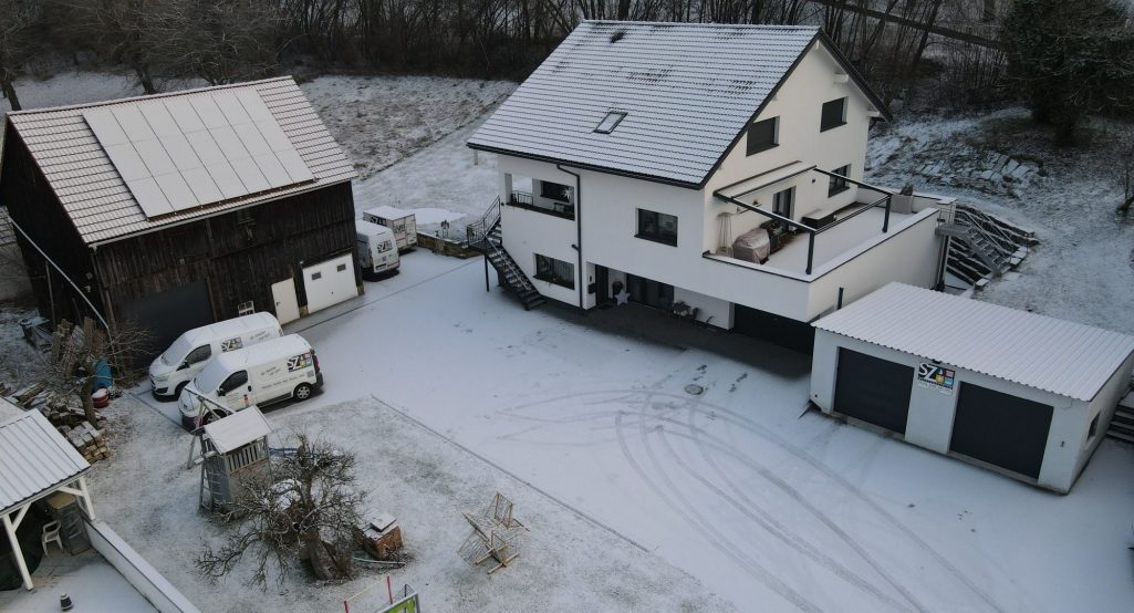 Gebäude SZ Sanitär-Heizung Schneebedeckte Landschaft mit einem Haus und Garagen, umgeben von Bäumen.