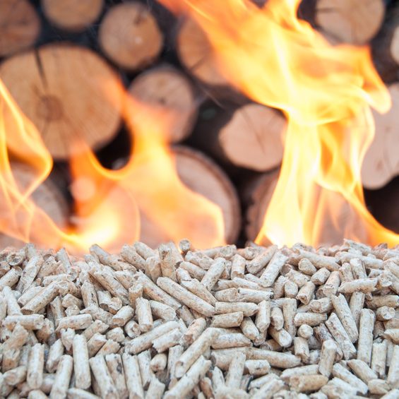 Brennholzpellets vor einem lodernden Feuer, im Hintergrund Holzstapel.